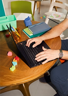Ein Mann arbeitet mit einem Laptop an einem Familientisch, auf dem Spielzeug und Büromaterial liegen.
